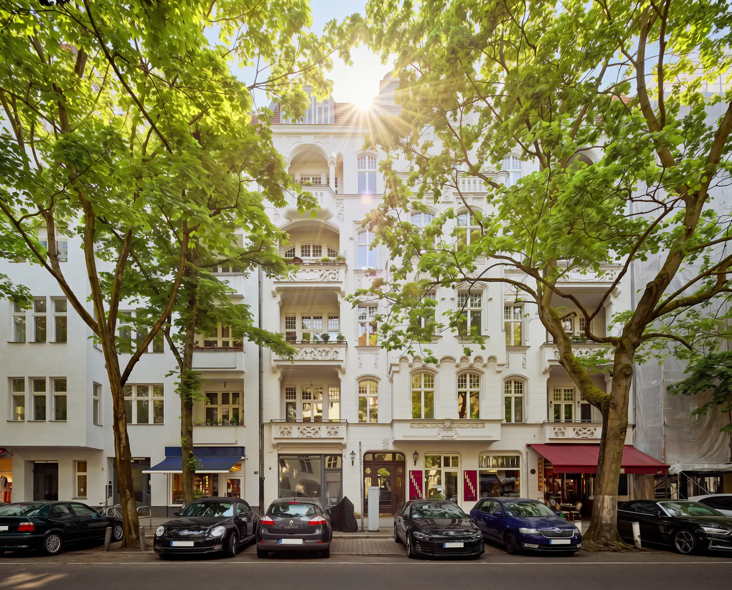 Street view of white apartment buildings with balconies, framed by green trees and parked cars. Sun shining above.