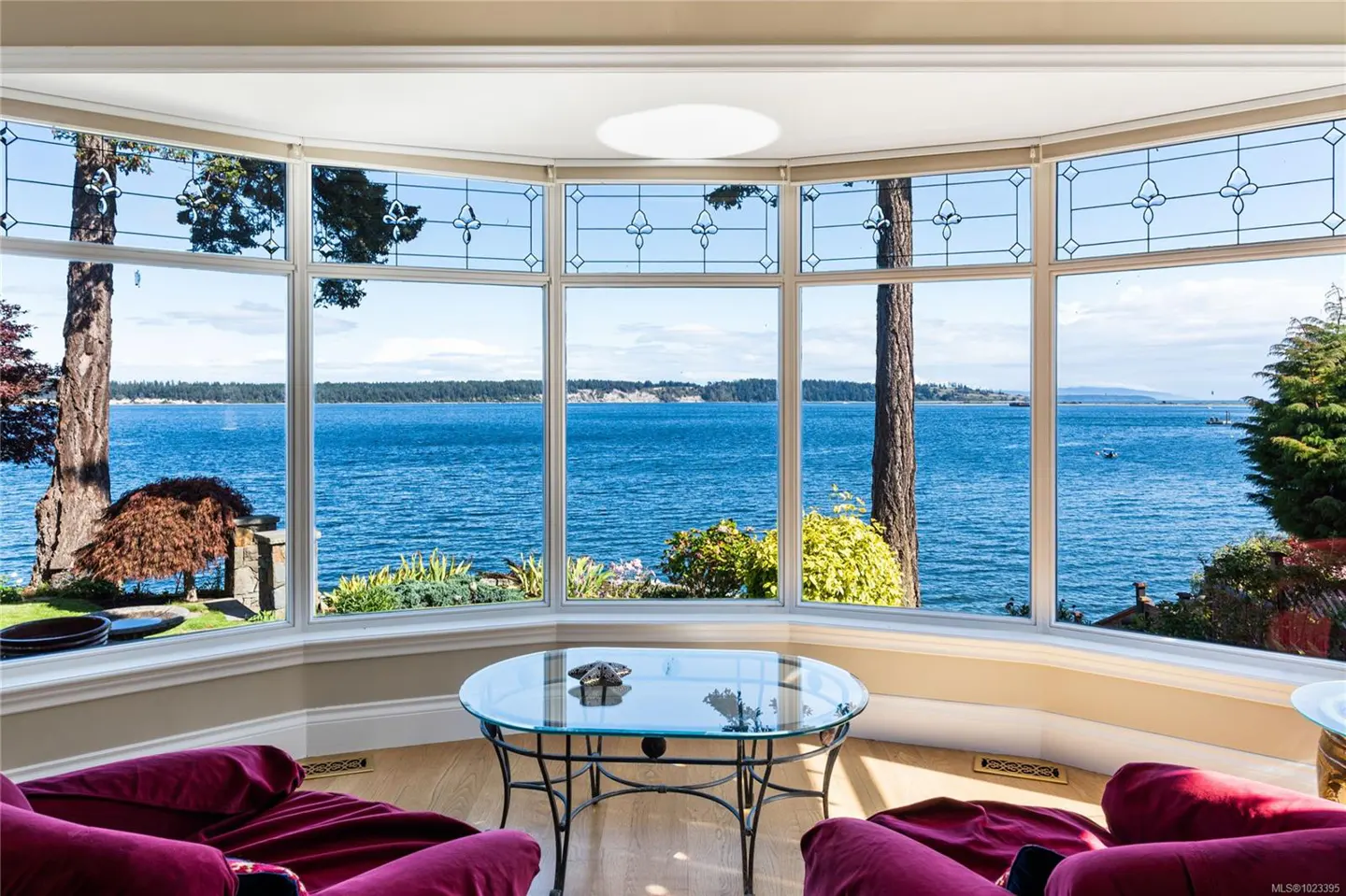 A bright living room with a bay window overlooking the ocean. Red chairs and a glass table are in the foreground.