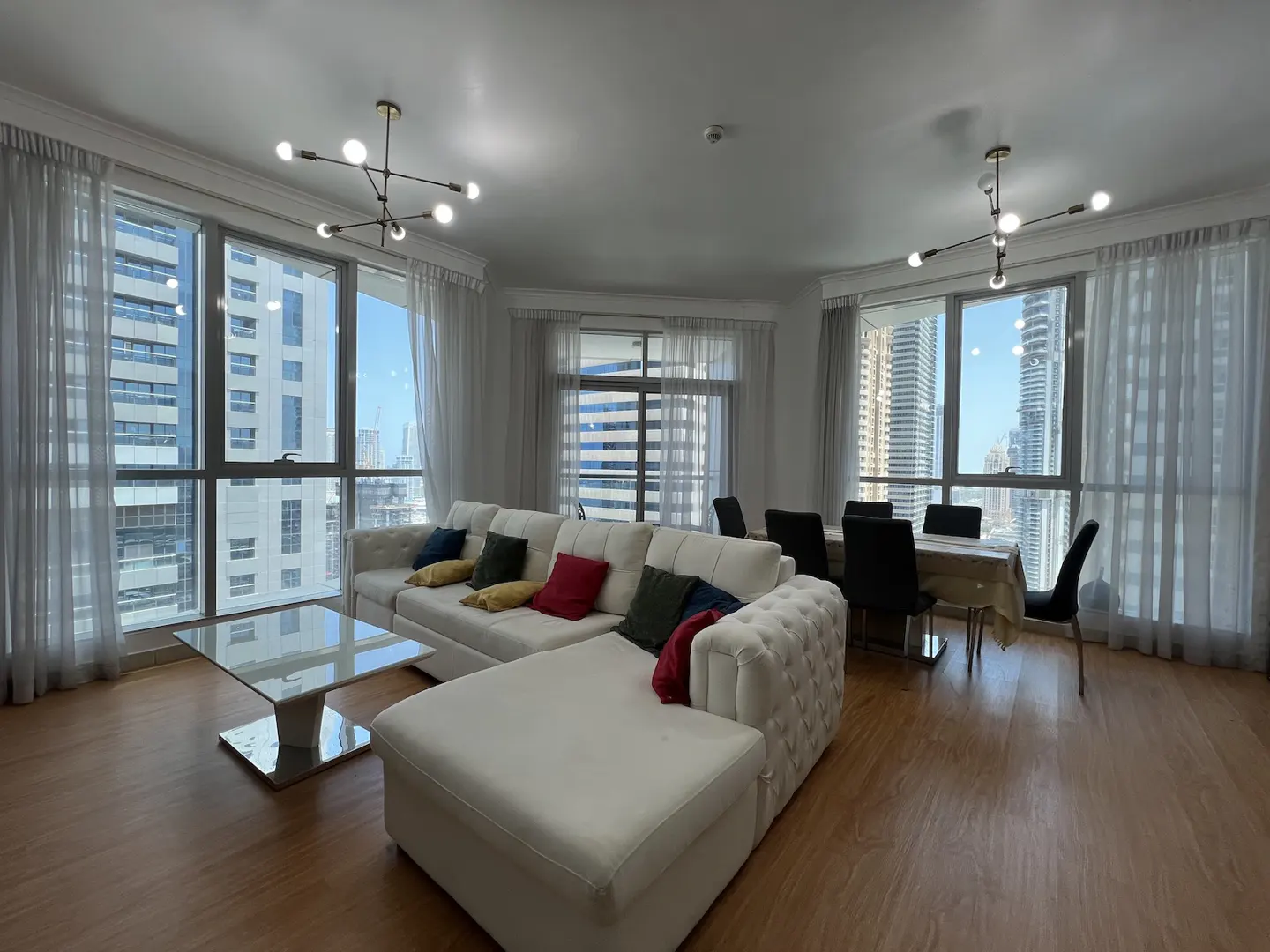 Bright living room with a white sectional sofa, colorful pillows, and a glass coffee table. Large windows offer a city view. Dining table with black chairs in the background.