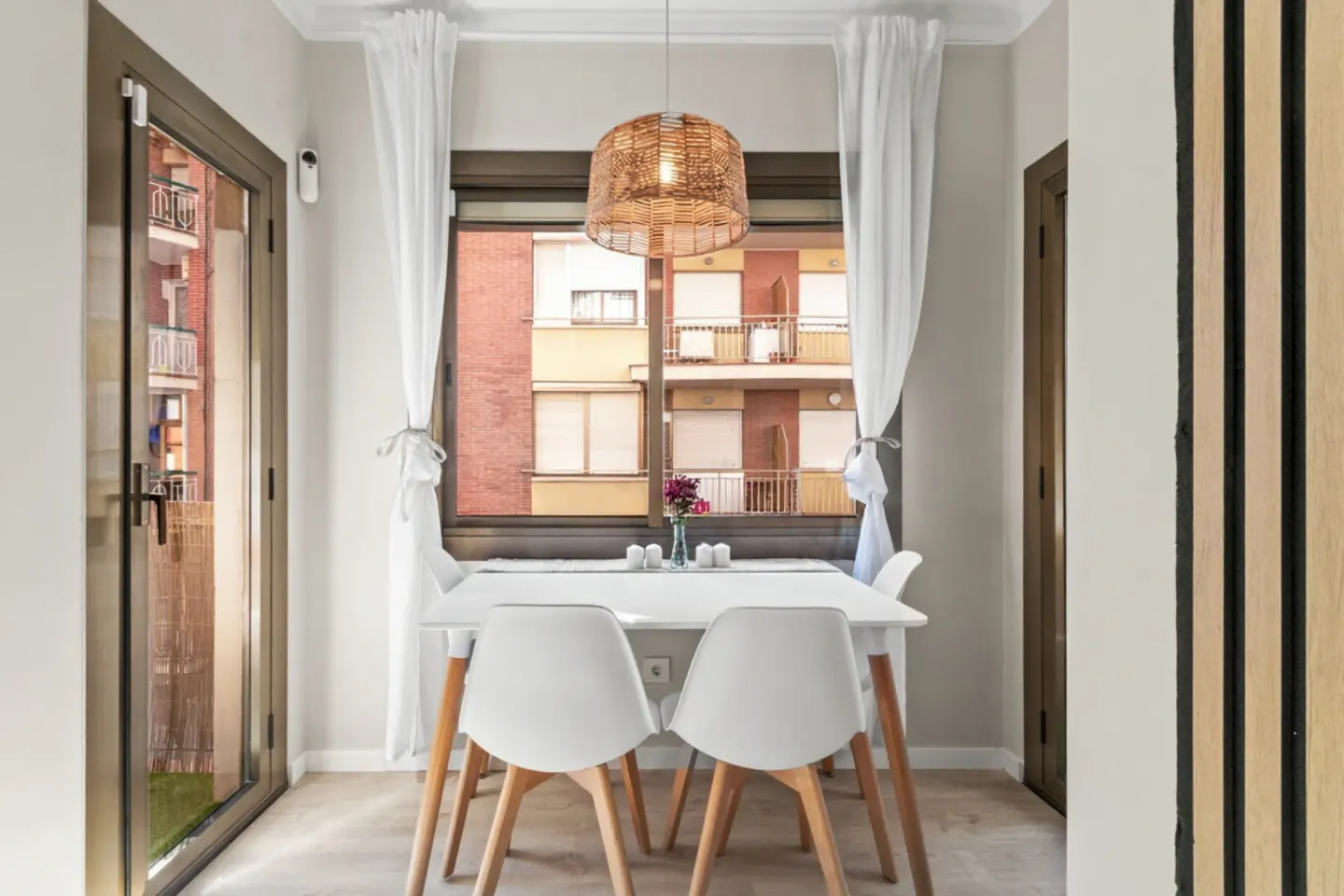 Bright dining area with a white table, chairs, and a woven pendant light. A window with white curtains overlooks a building.