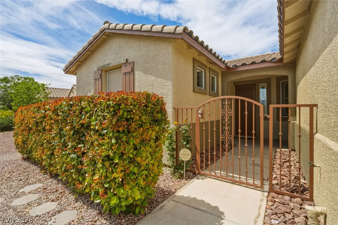 Tan stucco house with a brown tile roof, a brown gate, and a green hedge under a blue sky.