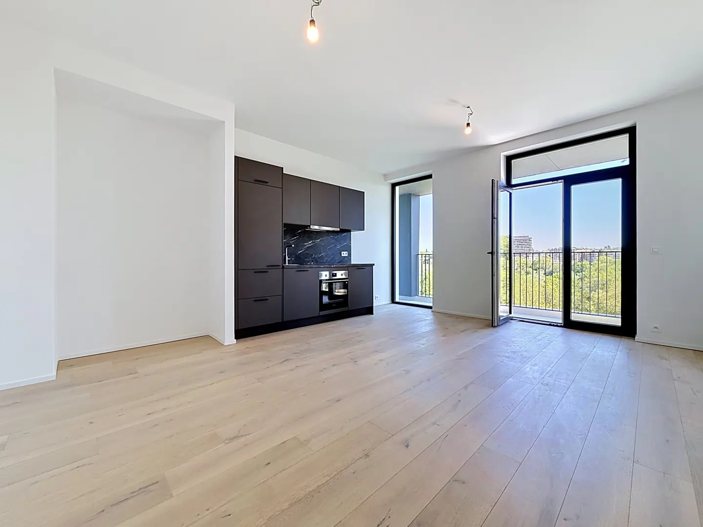 Bright, modern apartment with light wood floors, white walls, and a black kitchen. Balcony doors open to a city view.
