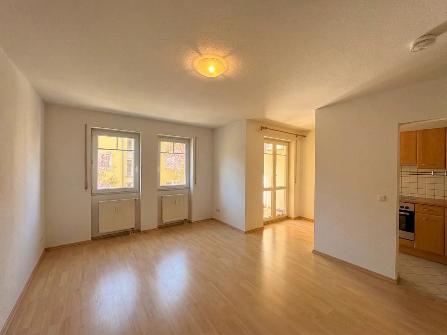 Bright, empty room with wood floors, white walls, two windows, and a door to a balcony. A kitchen is visible in the background.