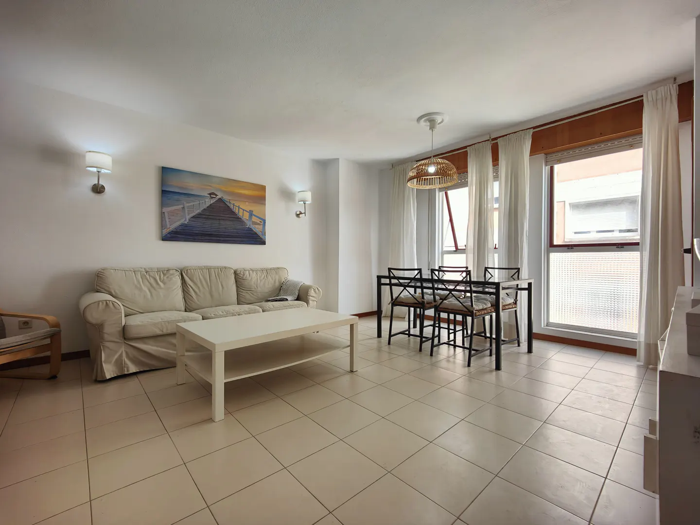 Bright living room with tile floors, a white sofa, a white coffee table, and a dining table with four chairs. A pier painting hangs on the wall.