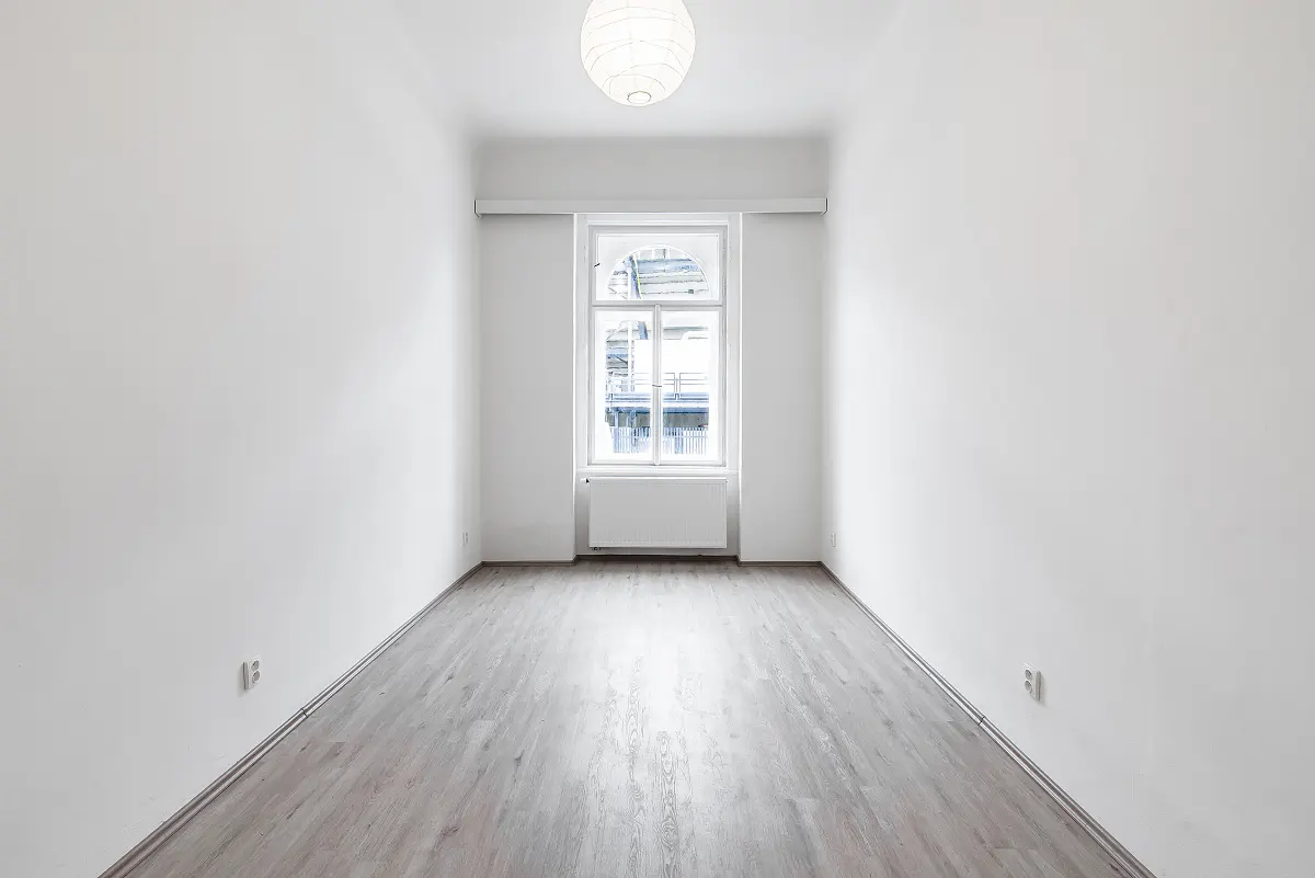 Empty room with white walls, light wood floor, and a white-framed window with a view of a building. A white globe light fixture hangs from the ceiling.