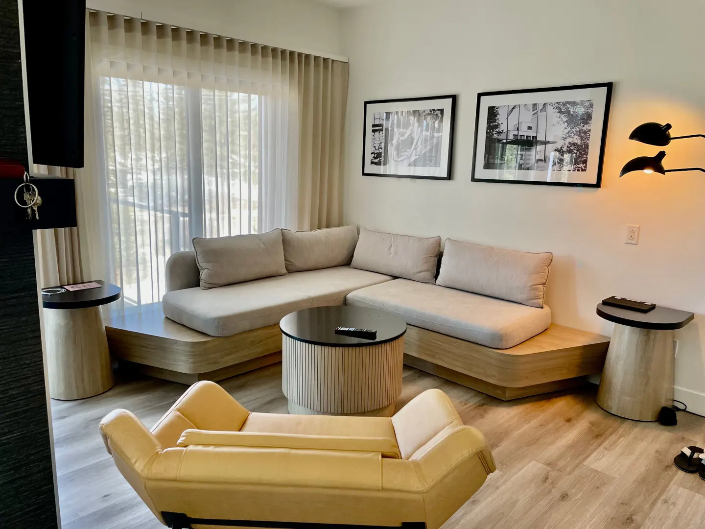 A modern living room with a beige sectional sofa, round coffee table, and a yellow lounge chair. Black and white framed art hangs on the wall.