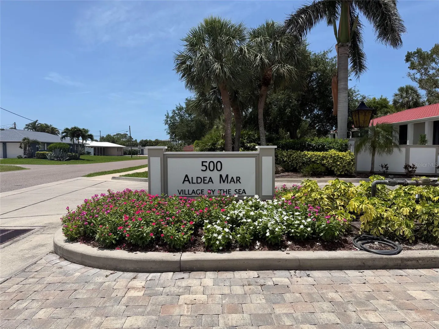 Outdoor sign reading "500 Aldea Mar Village by the Sea" with pink and white flowers in front, palm trees and blue sky in the background.