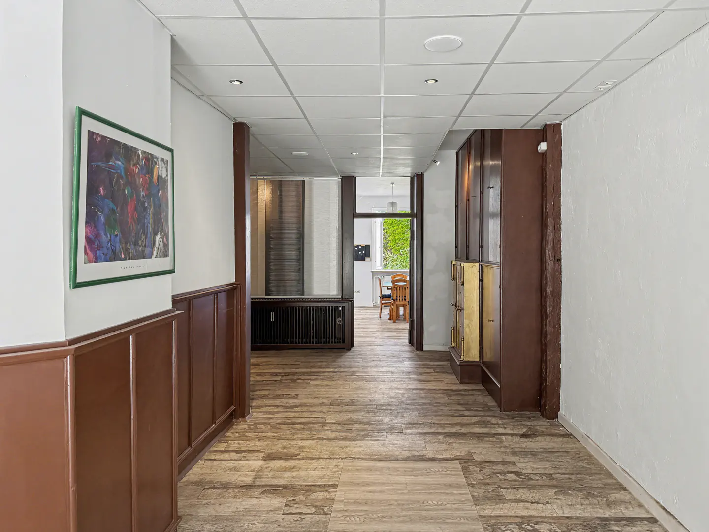 Hallway with wood-look floors, white walls, and brown wainscoting. A framed art print hangs on the left wall. A dining room is visible at the end.