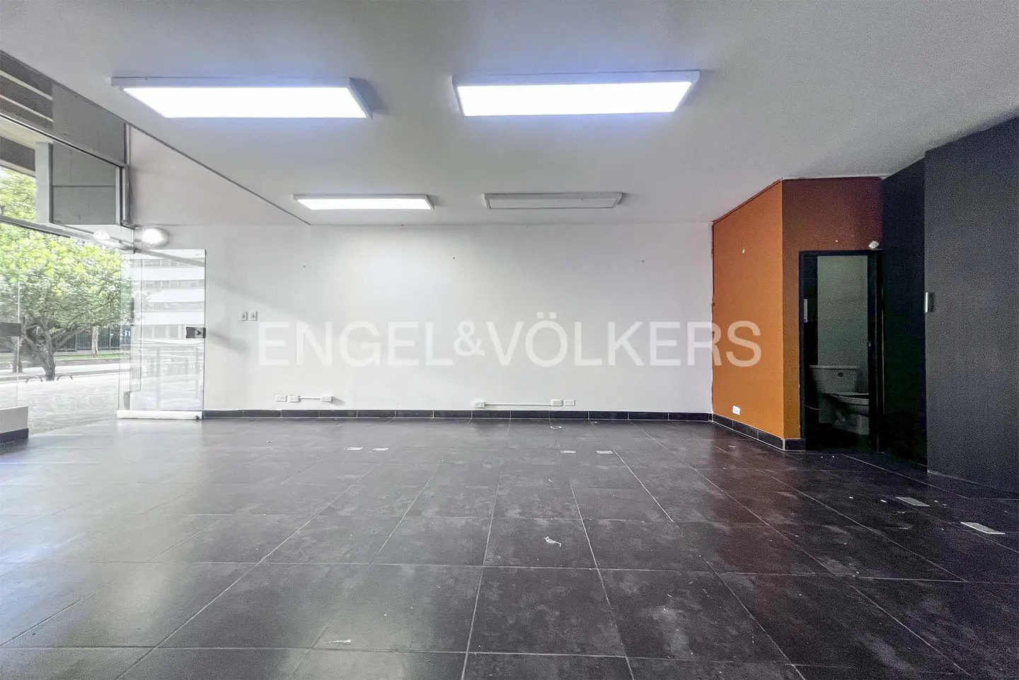Empty commercial space with black tile floor, white walls, and bright overhead lighting. Engel & Völkers logo on the wall.