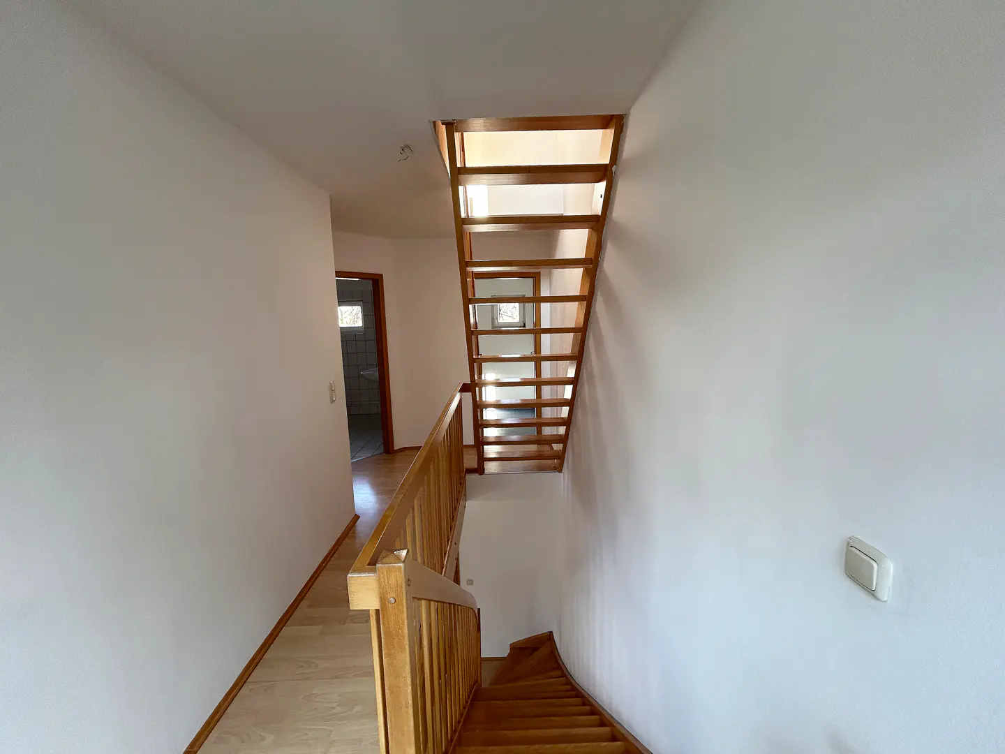 Bright hallway with white walls, light wood flooring, and an open wooden staircase leading to an upper floor.