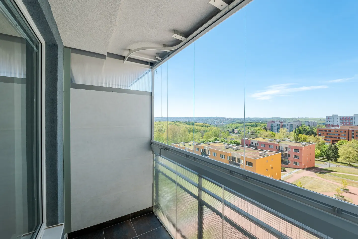 Apartment balcony with glass walls and metal railings overlooking a cityscape with green trees under a blue sky.