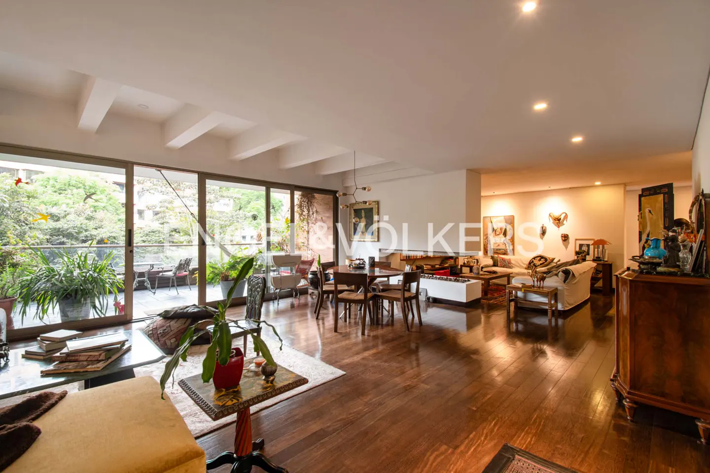 Wide shot of a bright, open-concept living and dining area with hardwood floors. A balcony with plants is visible through large glass doors.