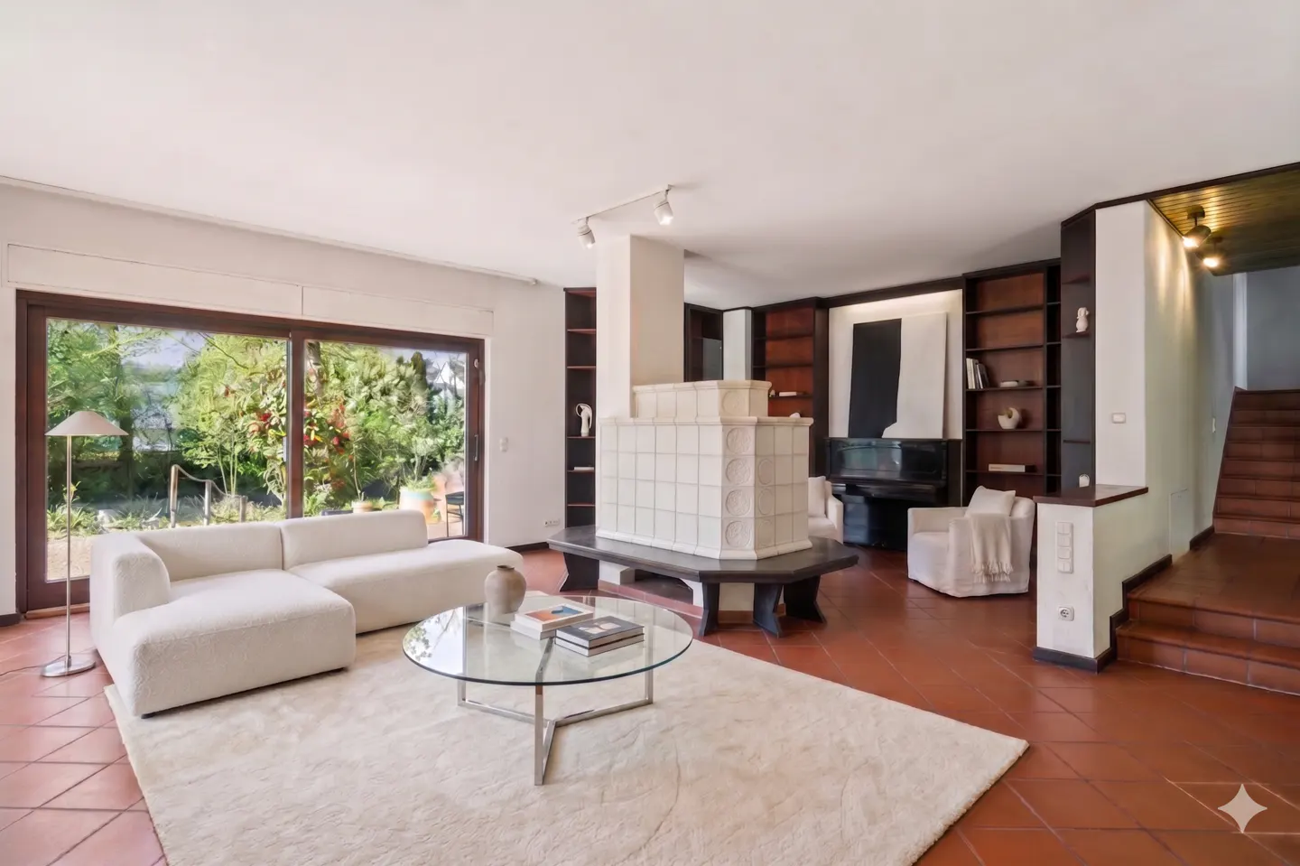 Bright living room with white couch, glass table, and tiled fireplace. Bookshelves and stairs are visible in the background.