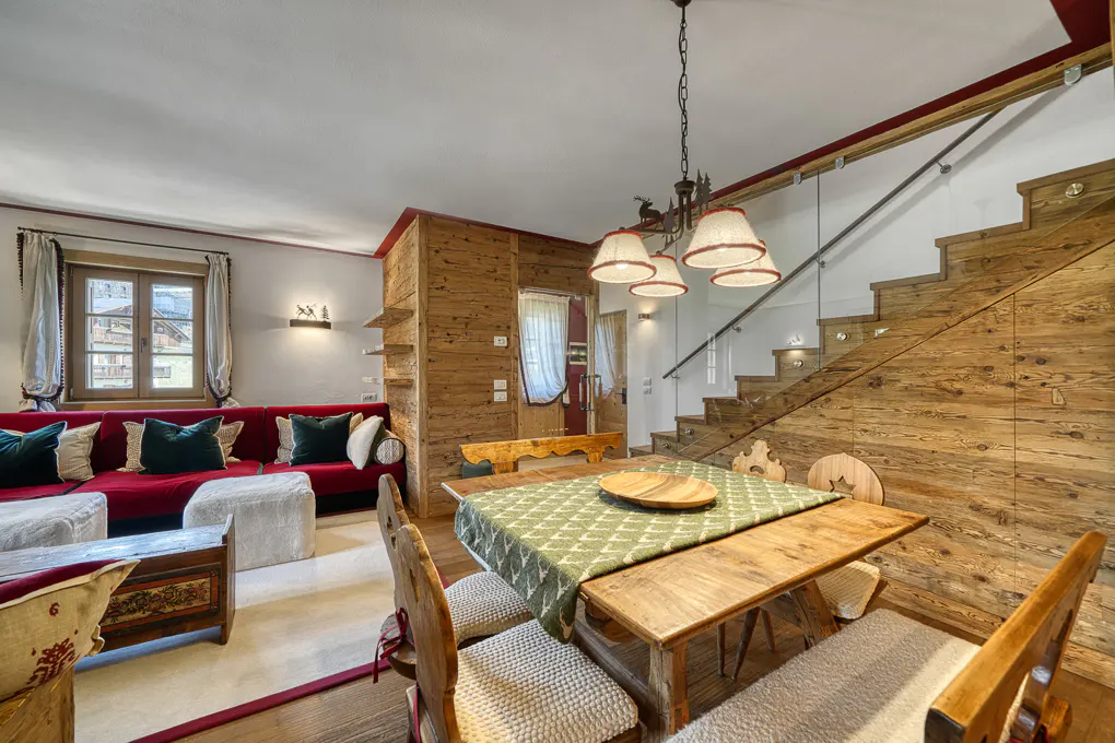 Cozy living room with wood walls, stairs, and dining table. Red sofa with green pillows sits near a window. Chandelier hangs above the table.