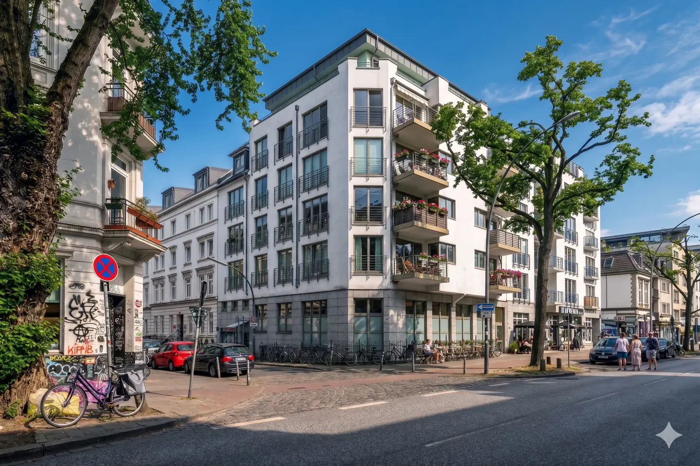 Street view of a modern white apartment building with balconies and flower boxes, trees, and parked bikes.
