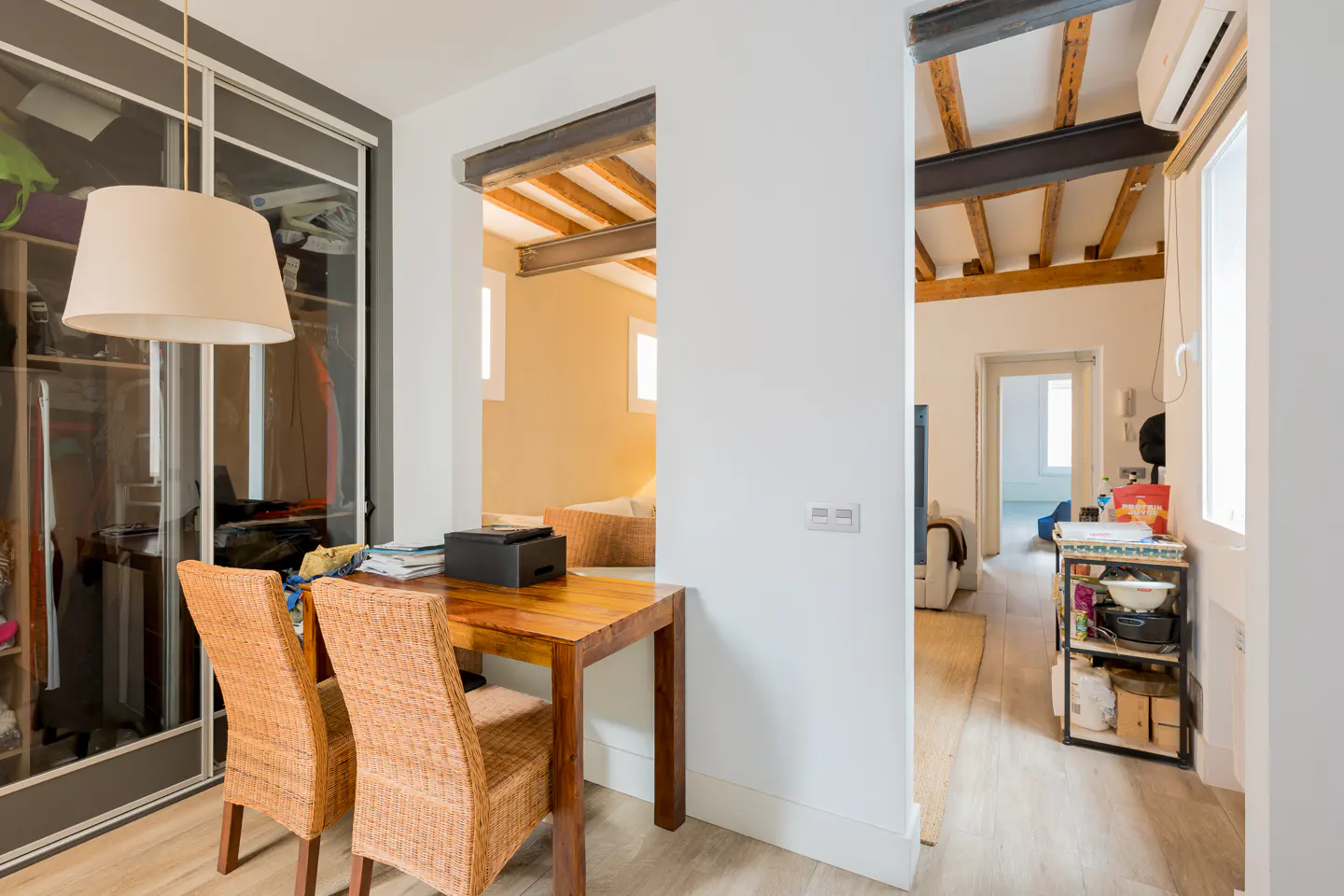 Interior view of a modern apartment with a wooden table, wicker chairs, and open doorways revealing rooms with exposed beams.