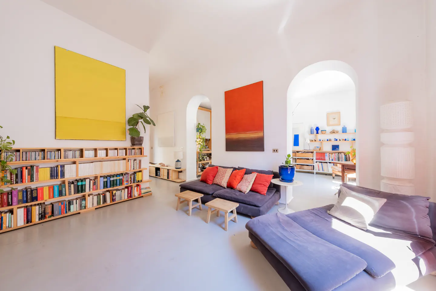 Bright living room with white walls, large yellow and red abstract art, a bookshelf, and dark sofas under arched doorways.