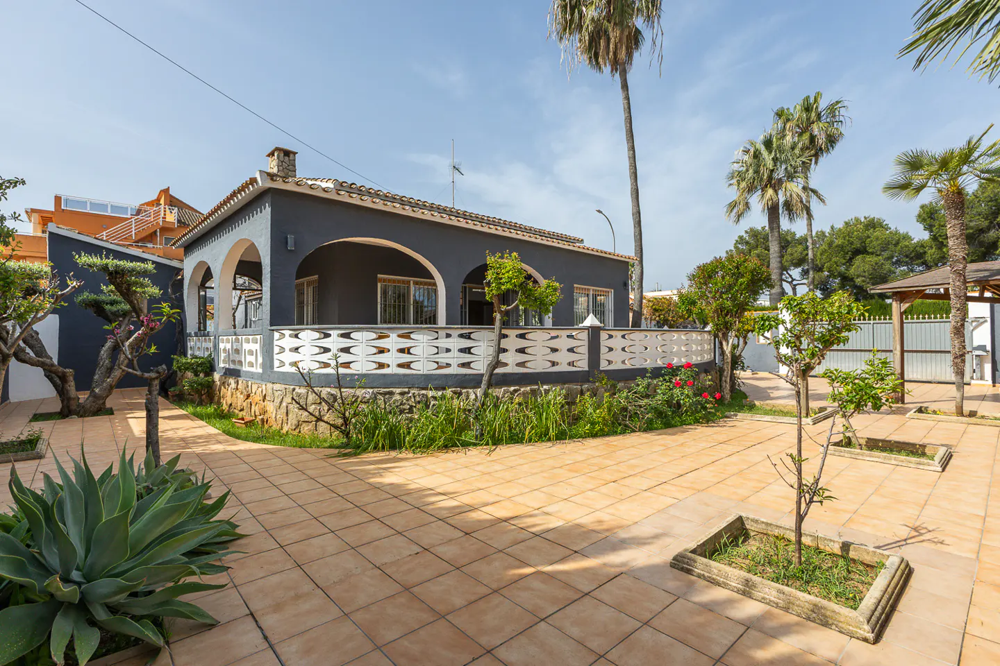 Exterior of a gray house with arched porch, white patterned fence, and tiled patio with palm trees.