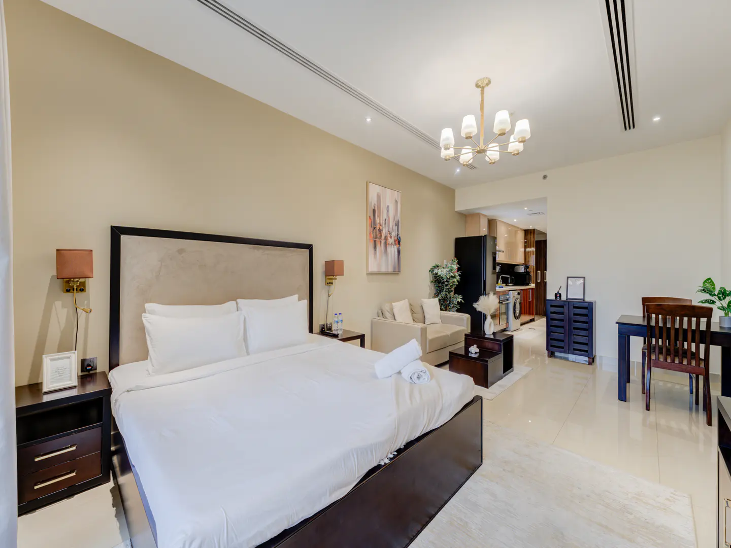 A modern studio apartment with a king bed, beige walls, and a small kitchen in the background.