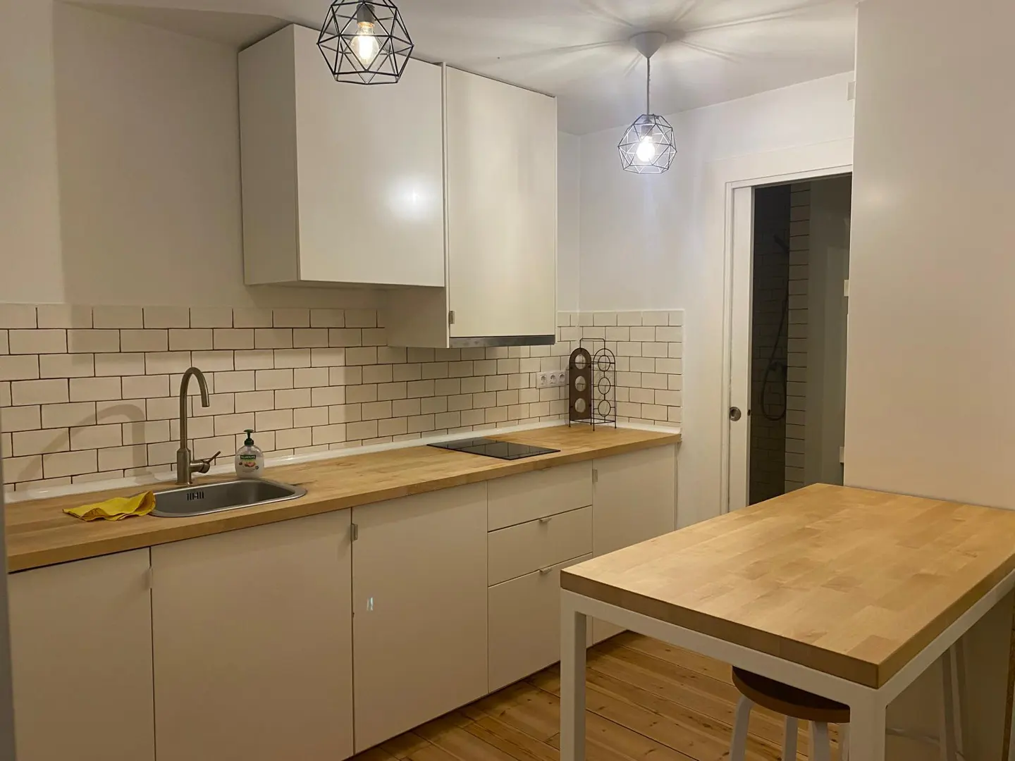 A bright kitchen with white cabinets, wood countertops, and geometric pendant lights. A table with a stool sits on a wood floor.
