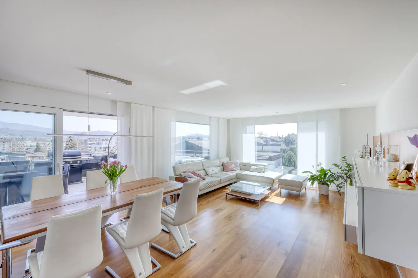 Bright, open-plan living and dining area with wood floors, white walls, and large windows. A wooden table with white chairs sits near a white sofa.