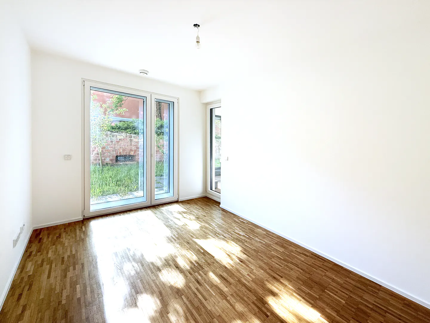 Bright, empty room with white walls and wood floors. Large glass doors lead to a garden. Sunlight streams in.