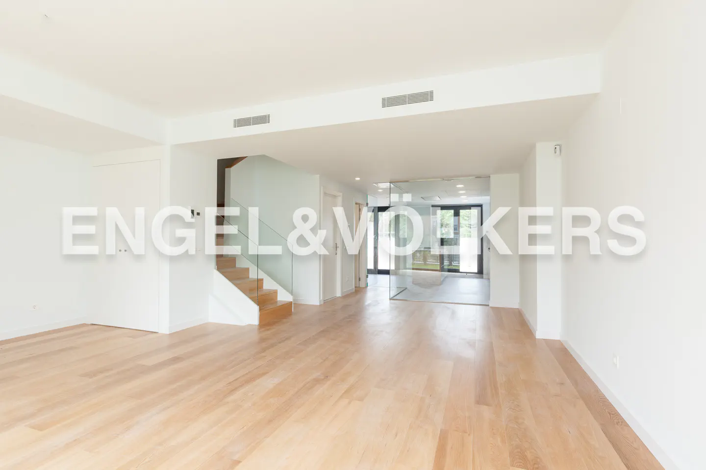 Bright, open-plan living space with light wood floors, white walls, and a staircase with glass railings. A glass-enclosed area is visible in the background.