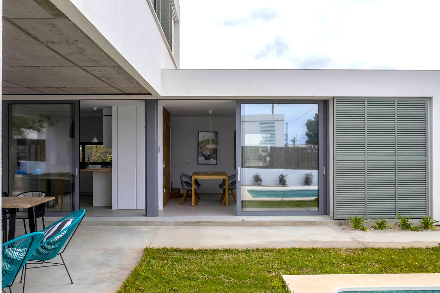 Modern home exterior with sliding glass doors leading to a dining area and kitchen, plus a pool view. Turquoise chairs on the patio.