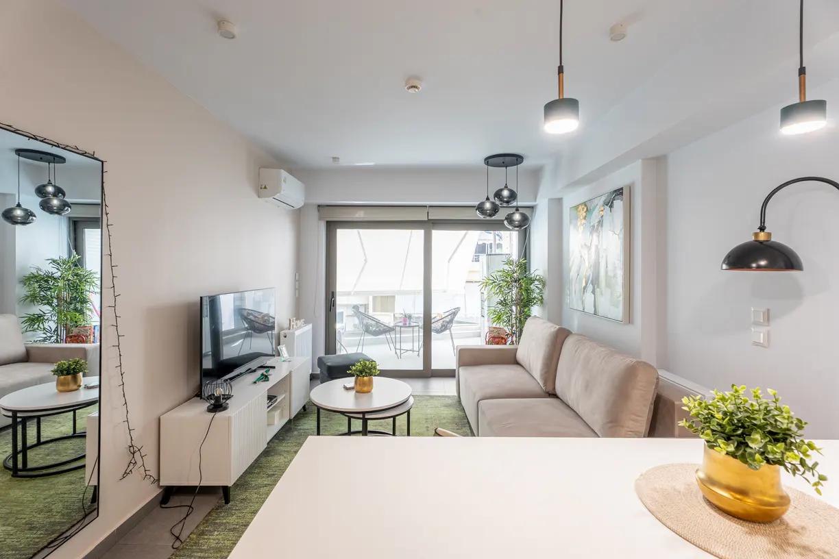 Modern living room with a beige sofa, white media console, TV, round coffee tables, and a balcony view with green accents.