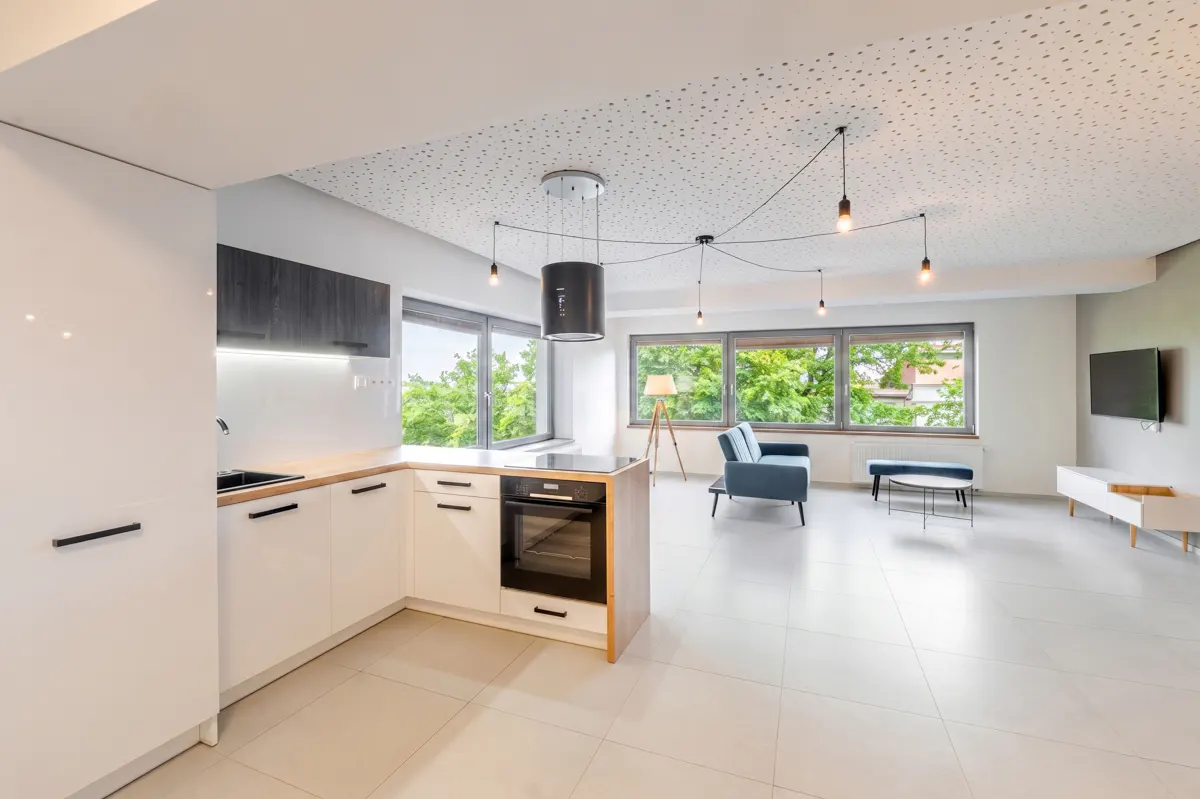 Bright, modern apartment with white tile floors. Kitchen with white cabinets and black hardware. Living area with blue sofa and large windows.