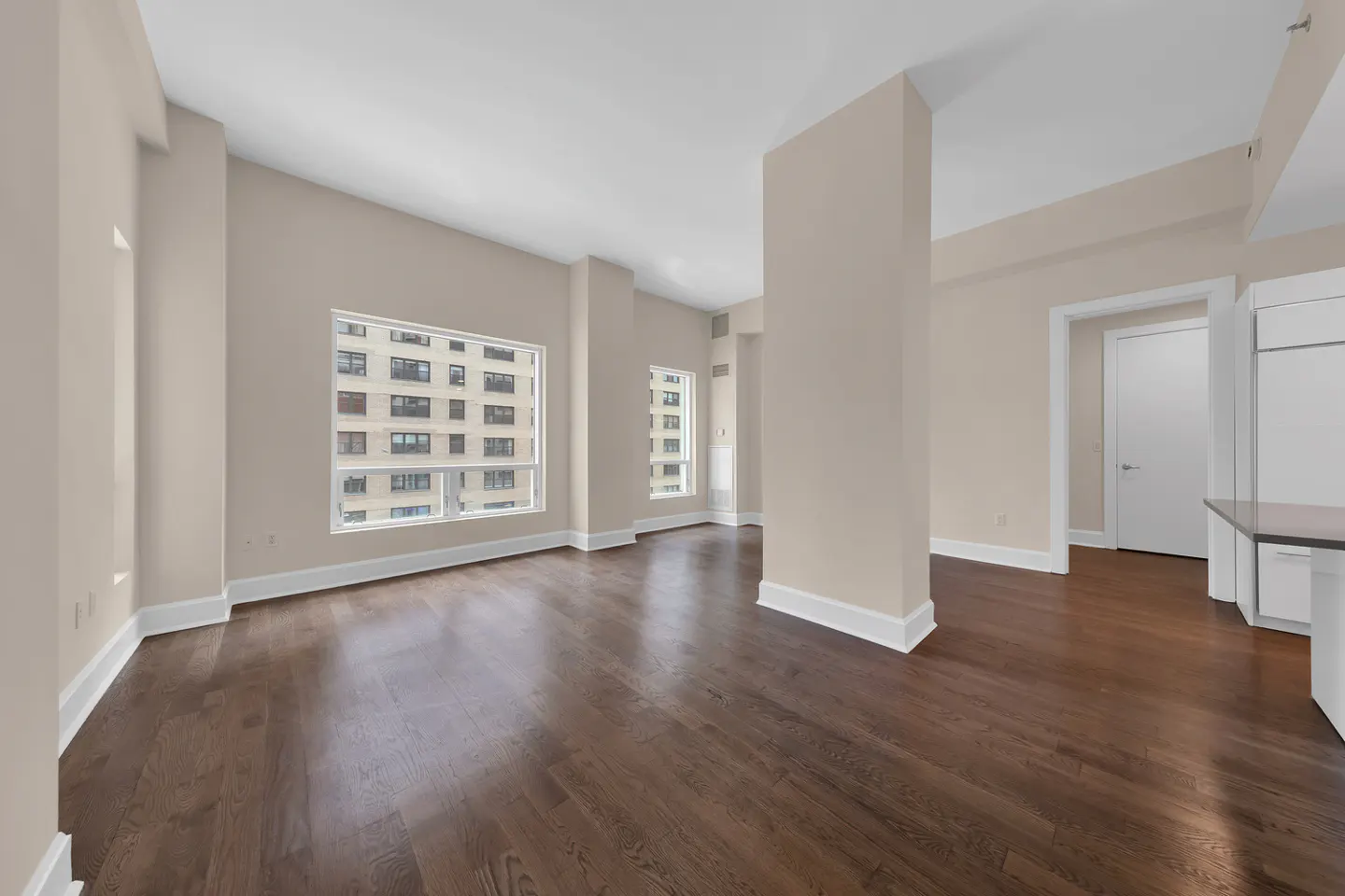 An empty living room with dark wood floors, beige walls, and large windows overlooking a city building.