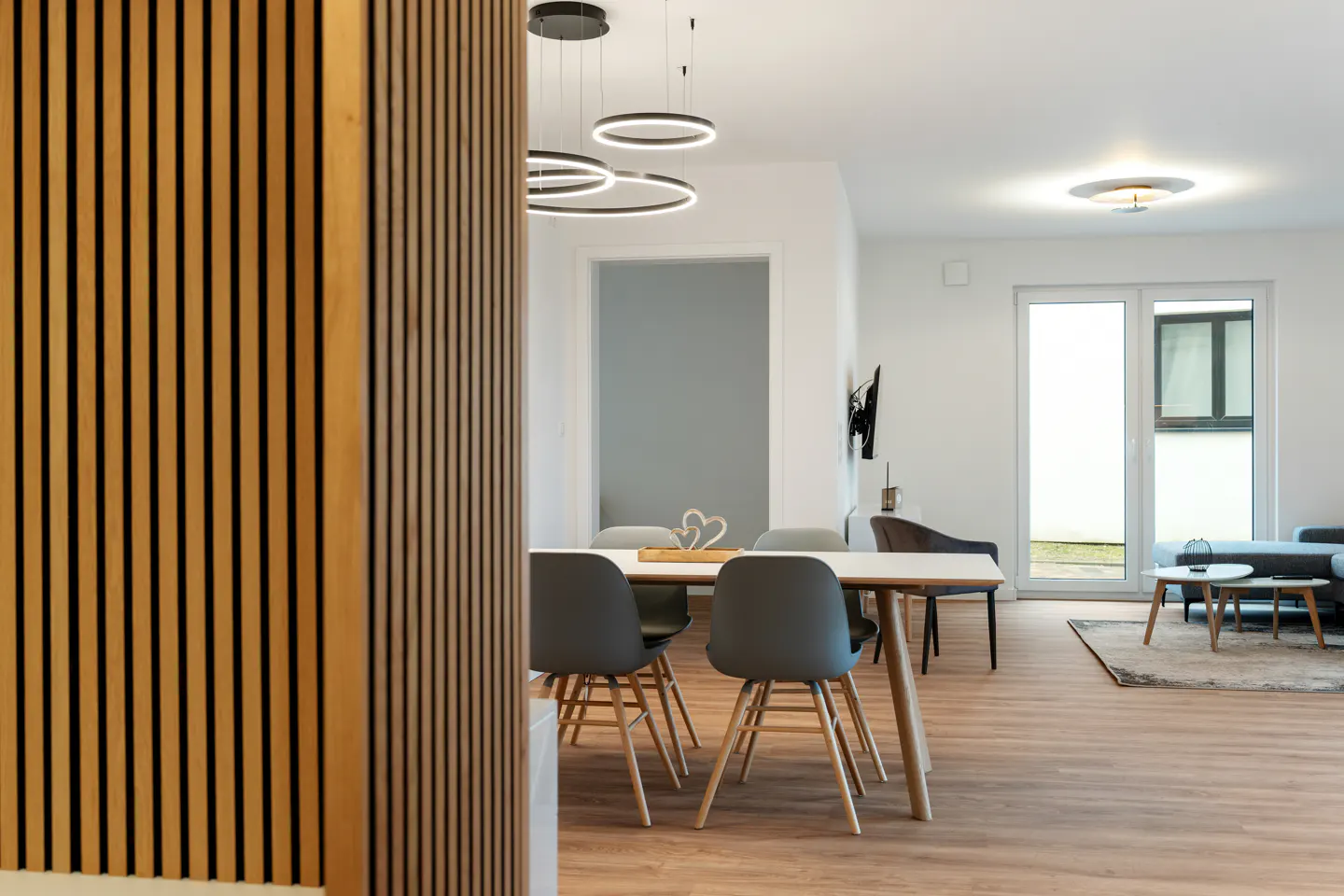 A bright, modern dining area with a white table, gray chairs, and wood floors. A wood slat wall is on the left. Circular lights hang above.