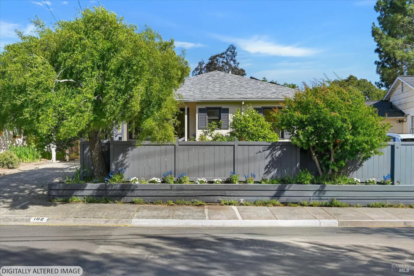 A single-story house with a gray roof is partially obscured by a gray fence and green trees. Blue and white flowers line the fence.