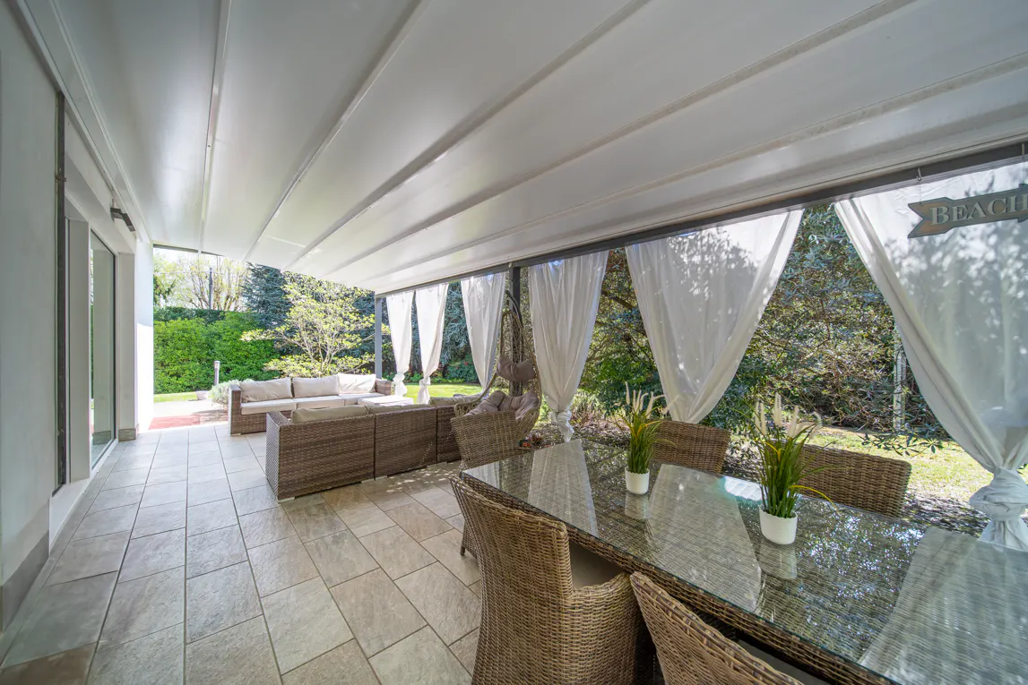 Covered patio with wicker furniture, glass table, and white curtains. A "Beach" sign hangs near the trees in the background.