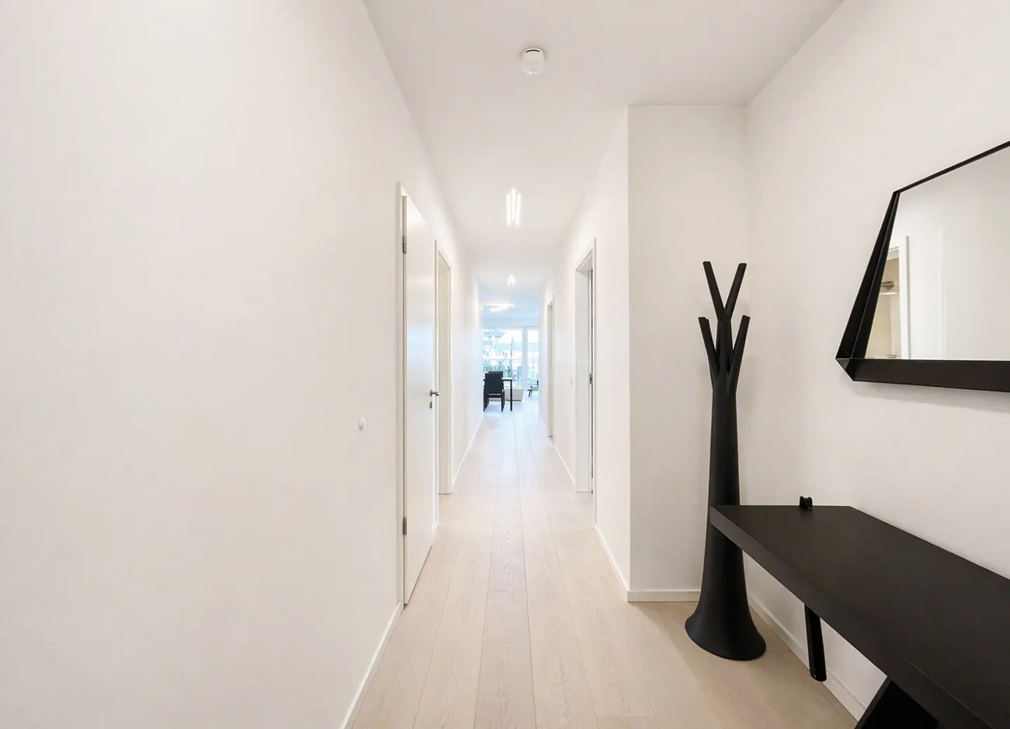 Hallway view with white walls, light wood floors, and black decor. A black coat rack, table, and geometric mirror accent the space. Doors line the hallway.