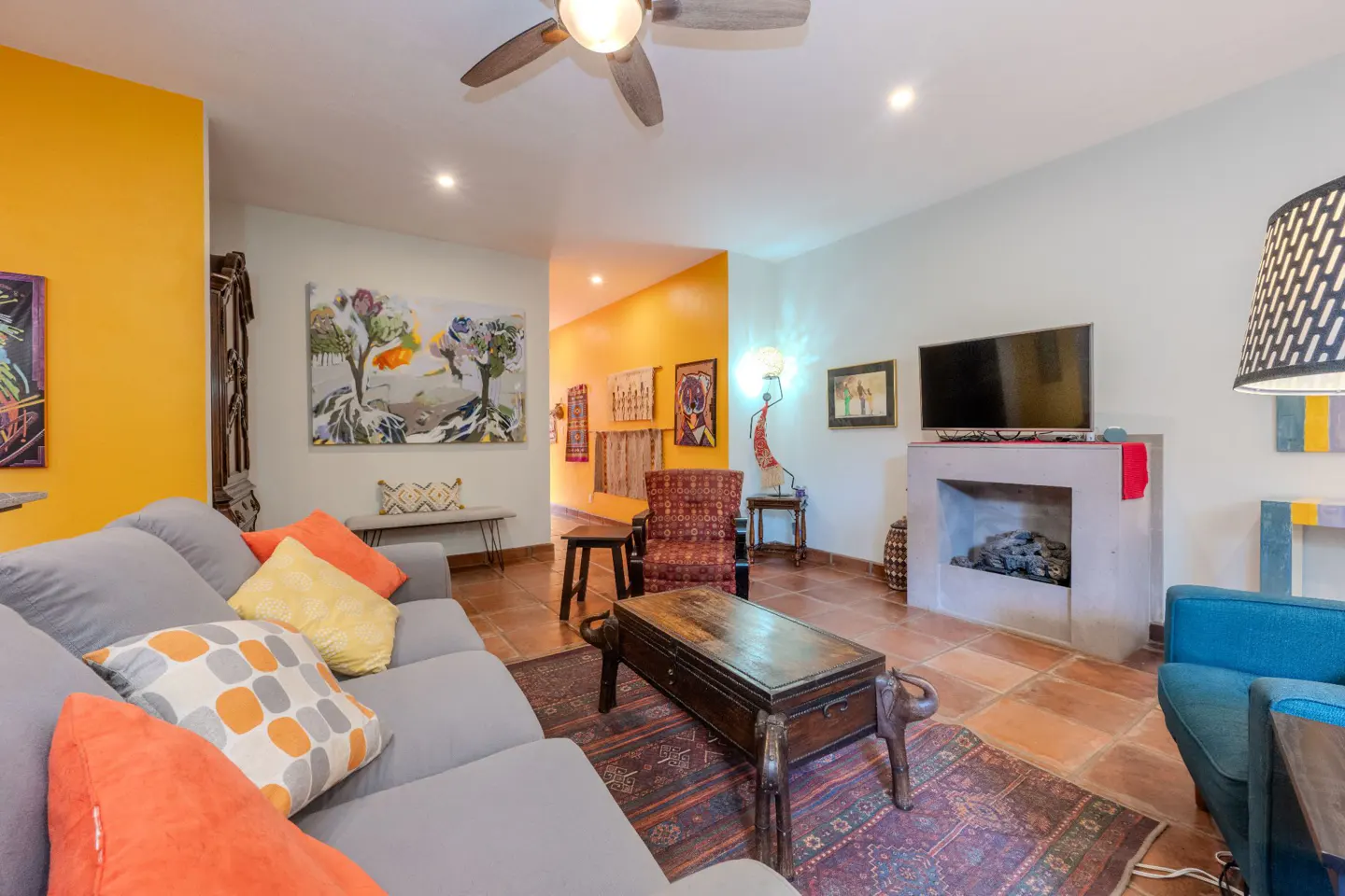 Bright living room with gray sofa, orange and yellow pillows, and a wooden coffee table with elephant legs. A fireplace and TV are on the right.
