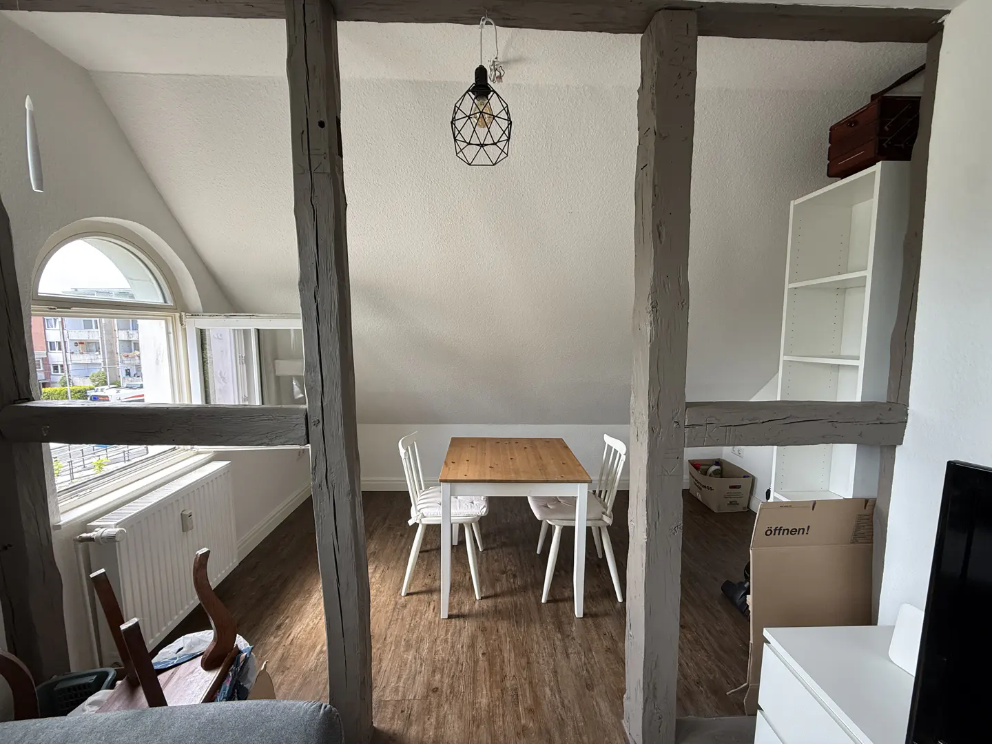 Attic room with wood-look floors, a dining table with two white chairs, exposed timber beams, and a window with a view.