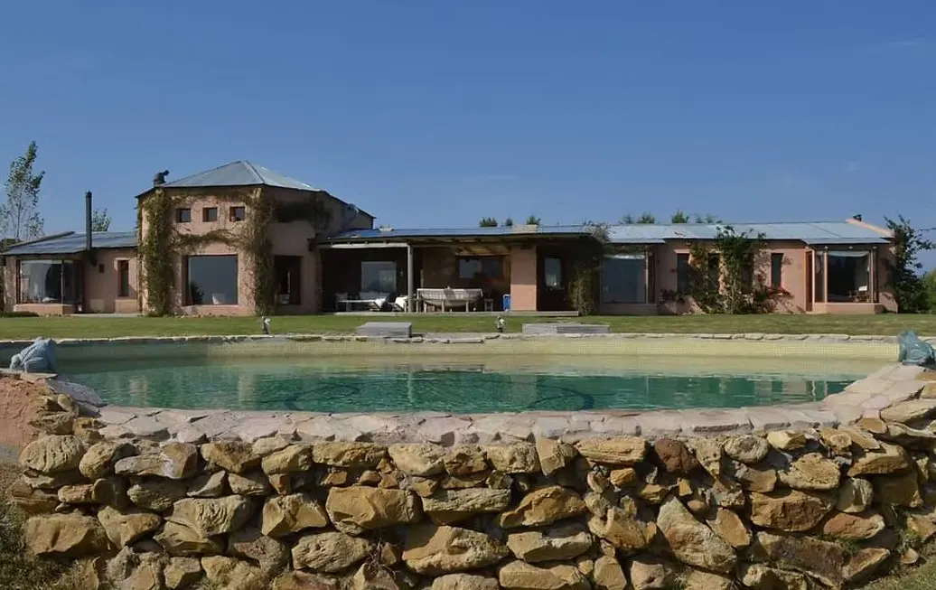 A pink house with a metal roof sits behind a stone-lined pool on a sunny day.