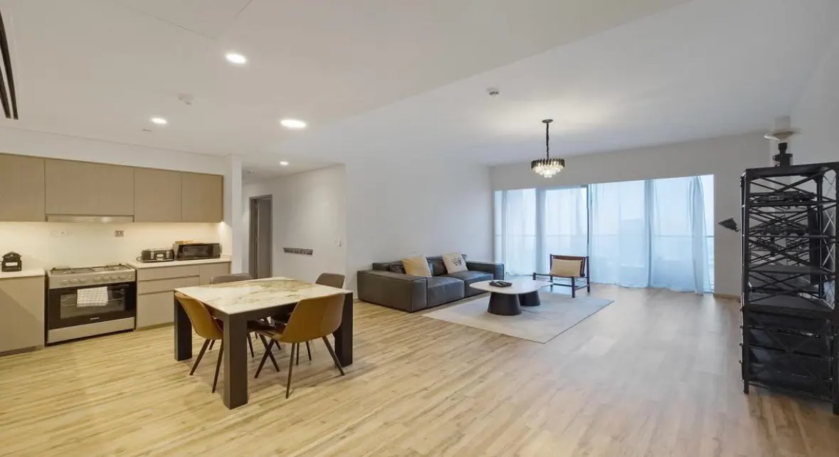 Open-concept apartment with kitchen, dining, and living areas. Beige cabinets, marble table, gray sofa, and city view.