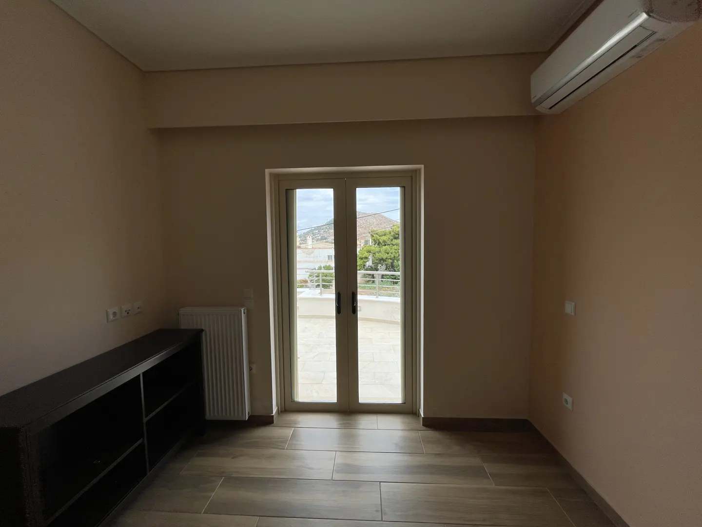 Empty room with beige walls, wood-look tile floor, and double doors to a balcony with a view. A black shelving unit is on the left.