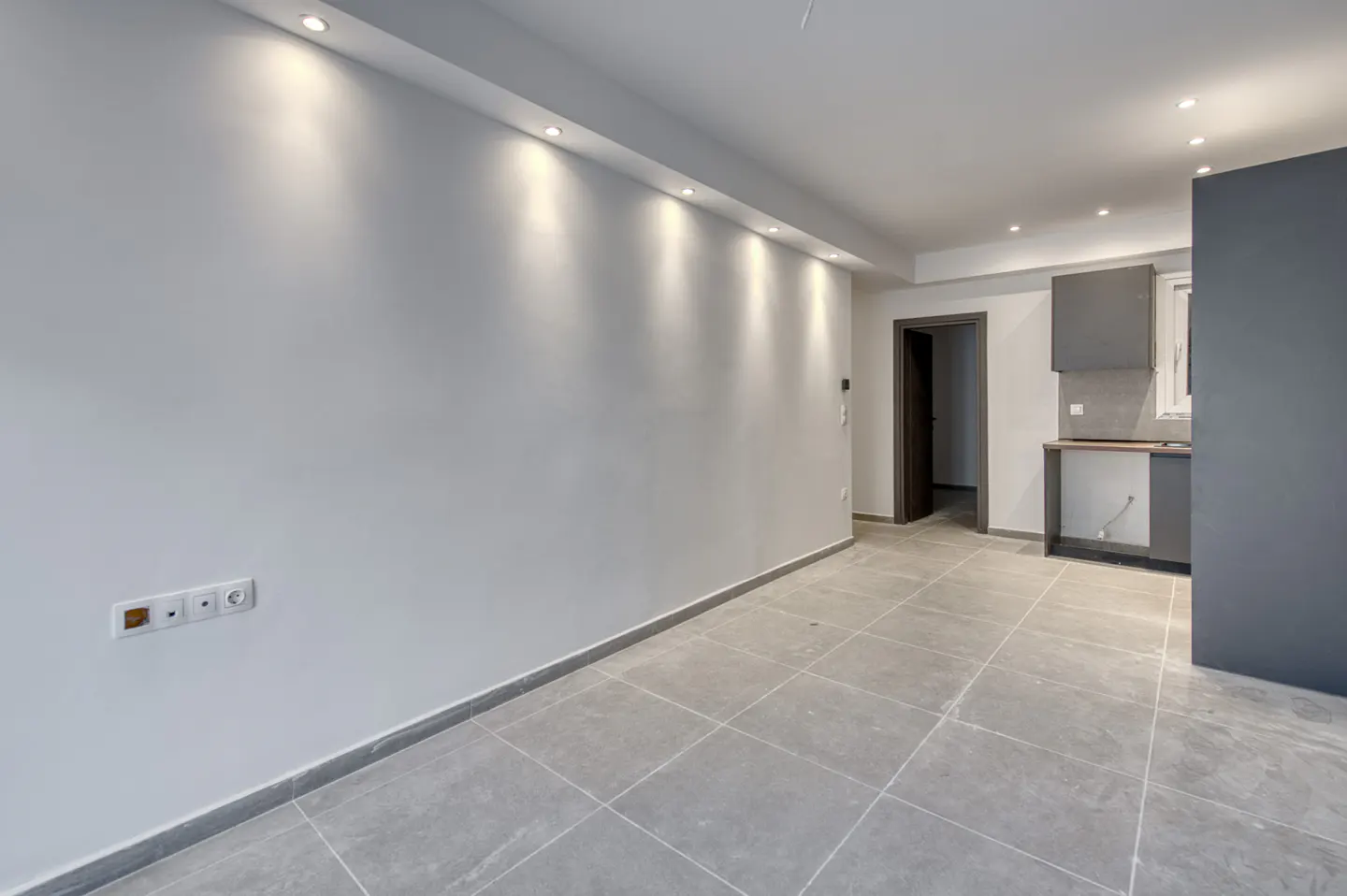Empty room with gray tile floor, white walls, and recessed lighting. A partial kitchen is visible in the background.
