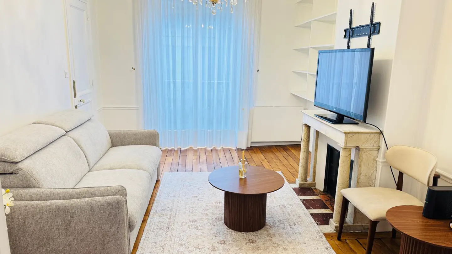 Living room with a gray sofa, round wood coffee table, TV on a marble fireplace, and a beige chair. The floor is wood and there is a light rug.