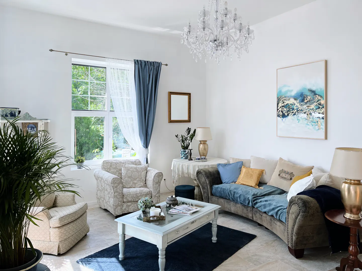 Bright living room with white walls, a chandelier, and a curved sofa with blue accents. Two armchairs and a coffee table sit on a dark rug.