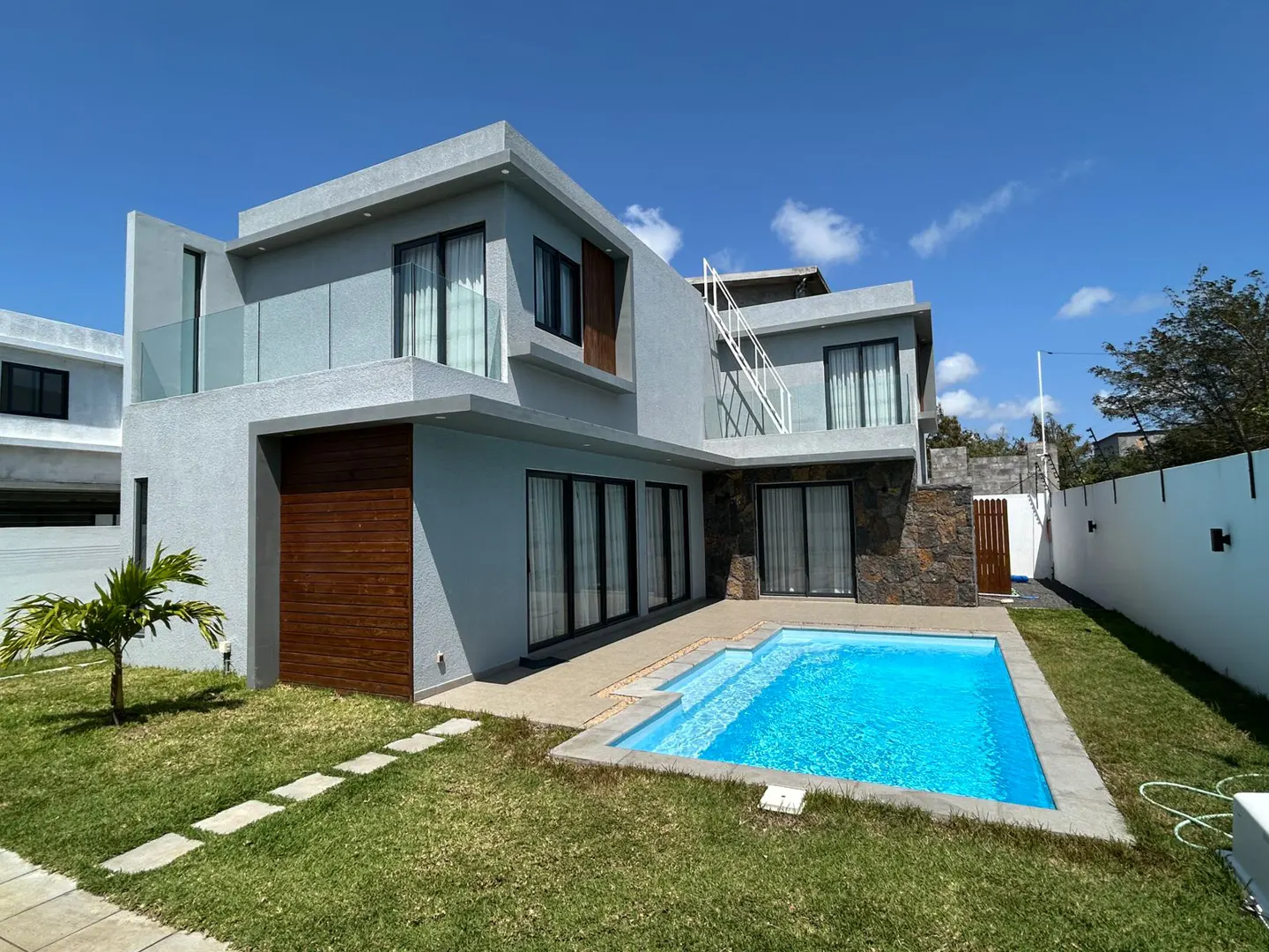 Modern two-story house with a blue swimming pool, green lawn, and a white fence under a blue sky.