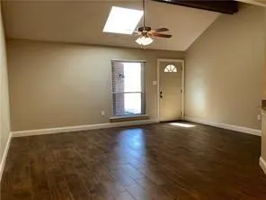 Empty room with dark wood-look tile floors, beige walls, a vaulted ceiling with a skylight, ceiling fan, and a front door.