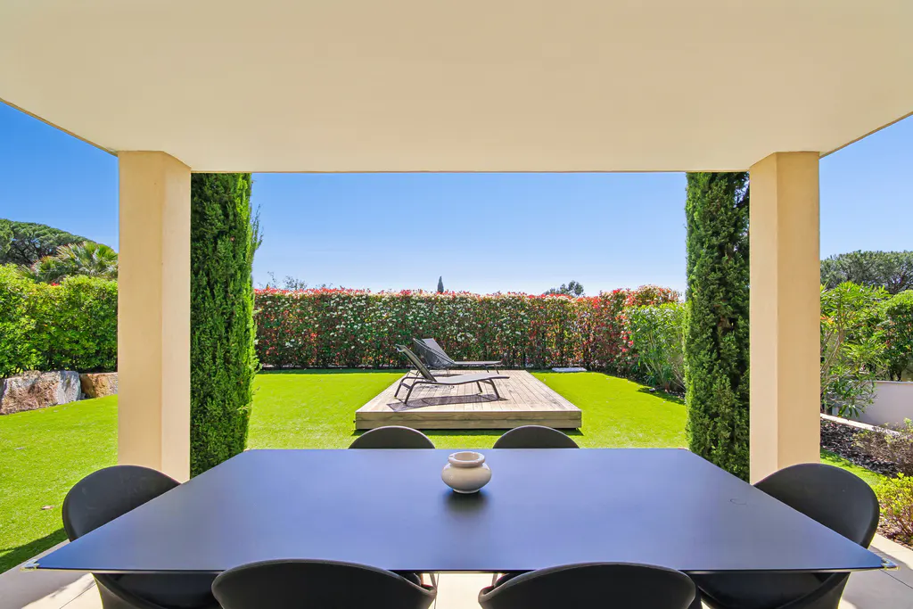 Covered patio with black table and chairs overlooking a green lawn, lounge chairs, and a hedge under a clear blue sky.