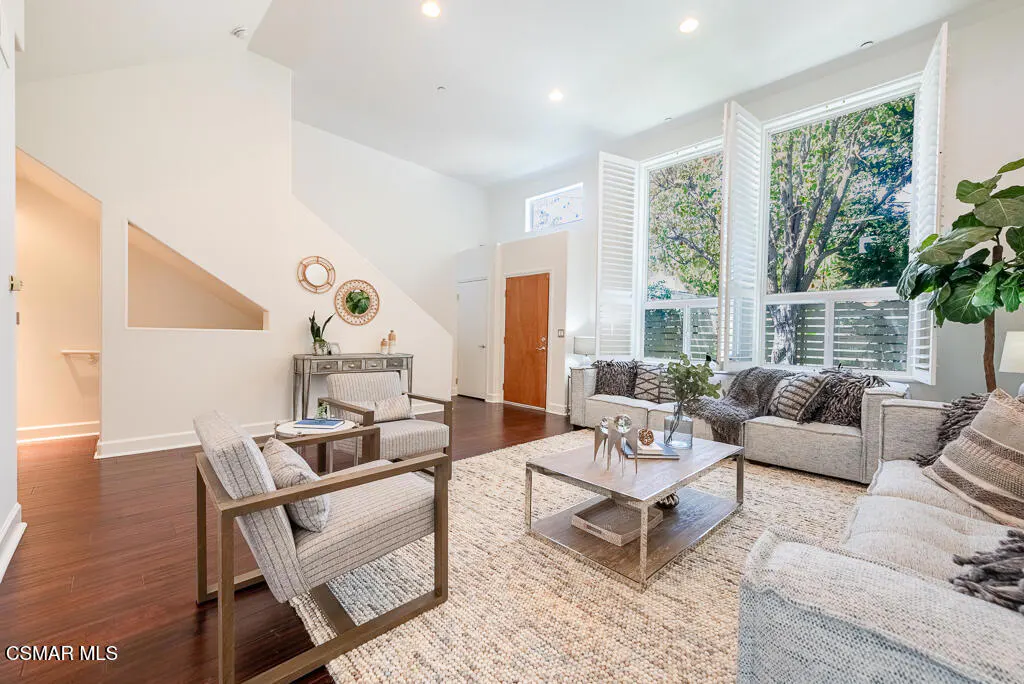 Bright living room with white walls, wood floors, and large windows with white shutters. Gray sofas and chairs surround a light-colored rug and coffee table.
