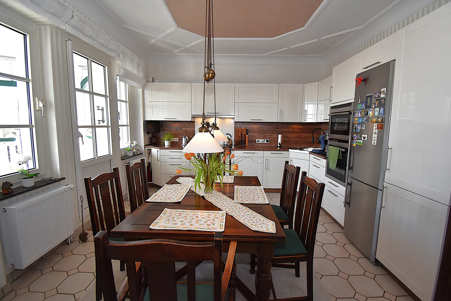 A bright kitchen with a dark wood table and chairs, white cabinets, and stainless steel appliances. A vase of orange tulips sits on the table.