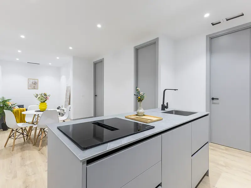 Modern kitchen with gray island, black cooktop, and sink. Open floor plan with dining table and white chairs. Light wood floors and gray doors.