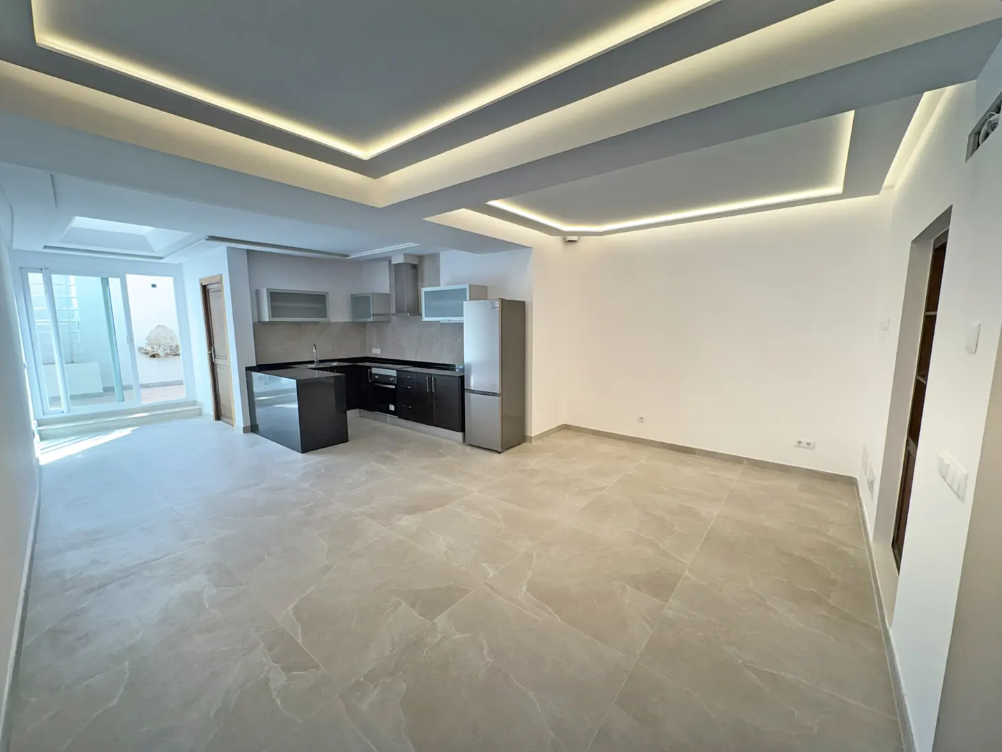 A bright, empty room with gray marble floors, a black kitchen island, and a stainless steel refrigerator.